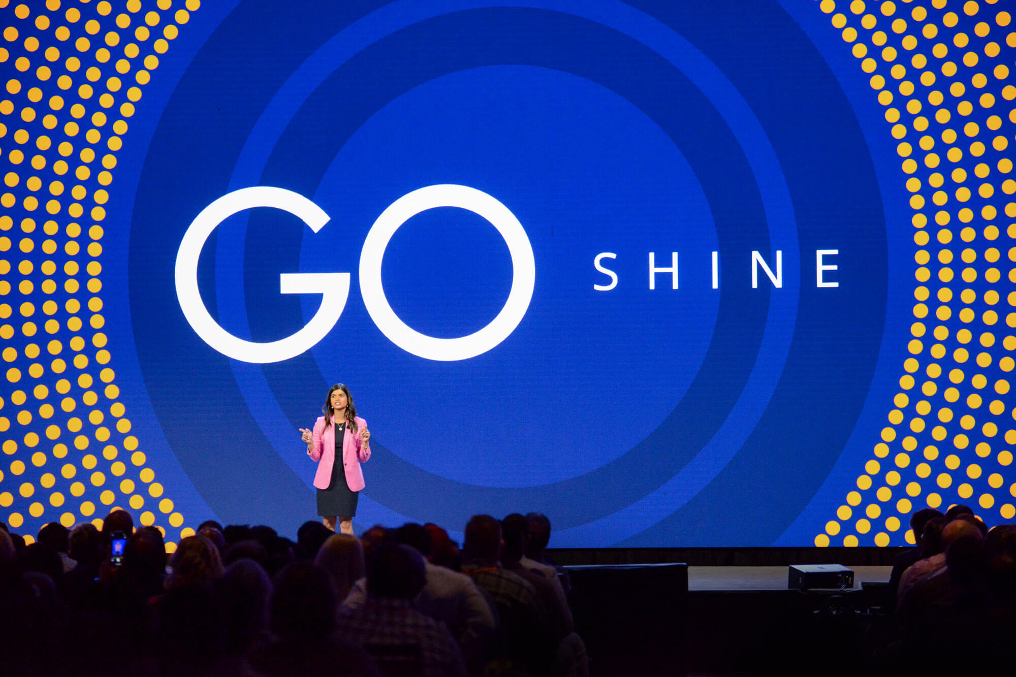 Woman presenting on stage with the words "Go Shine" on the screen behind her